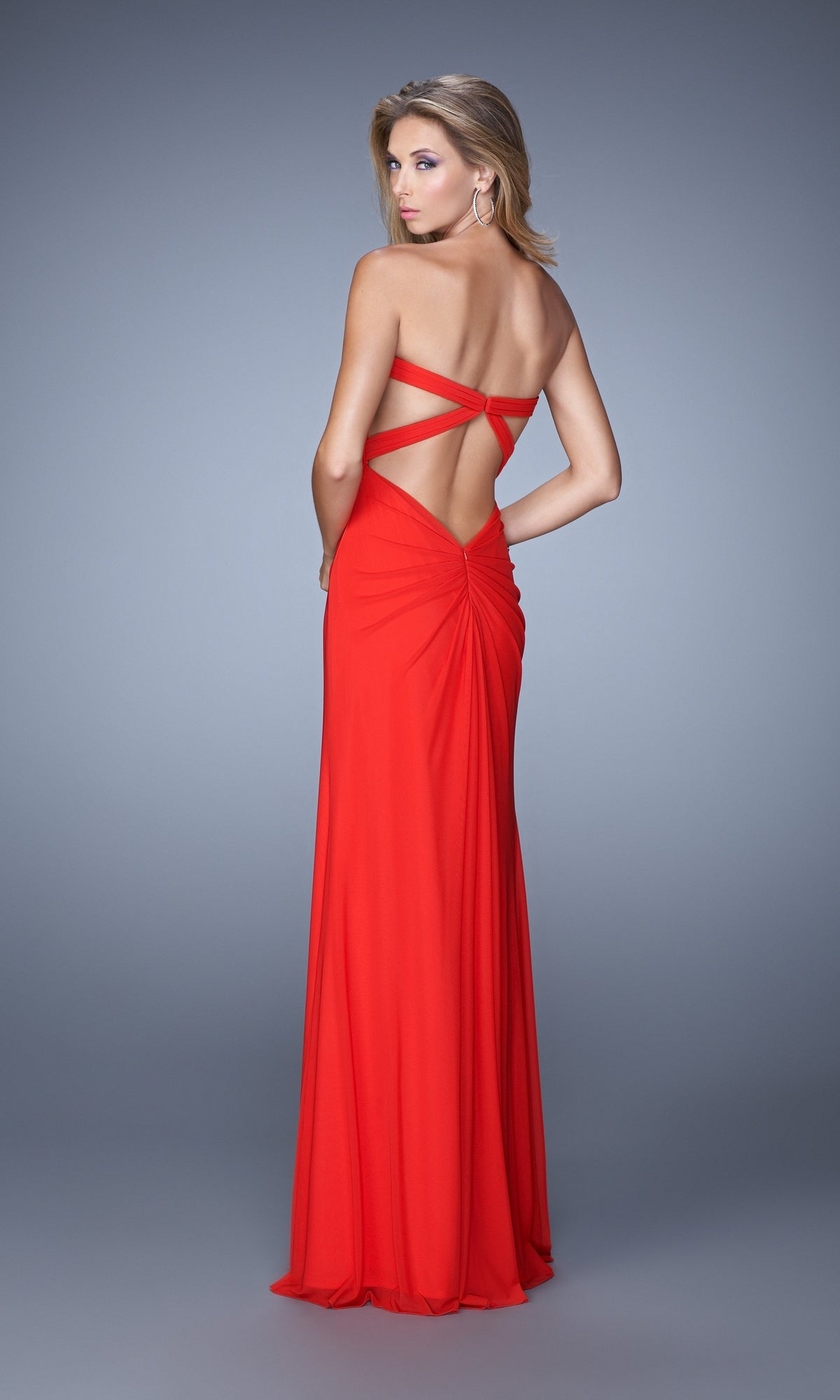 Woman wearing the La Femme Dress 21233, a floor-length red jersey net gown with crisscross straps and a strapless bodice, posing against a plain gray background.